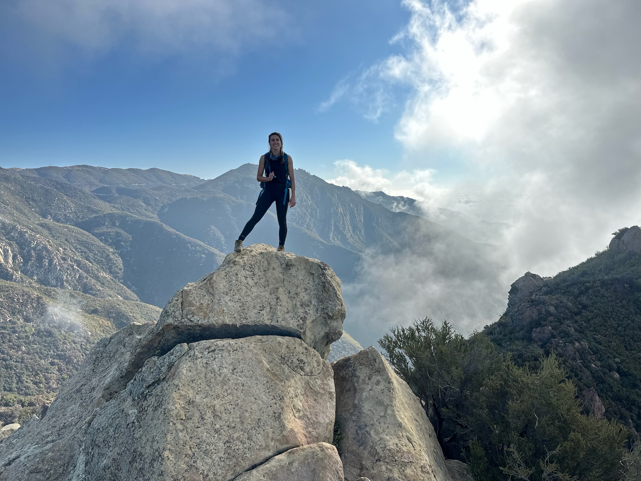 A woman stands on top of a rocky peak with blue sky and chapparal-covered mountains in the background. Blue sky is visible to the left, and foggy, dark clouds are rolling in from the right.